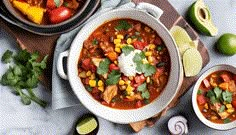 two bowls of chili and corn soup on a table with limes, cilantro, avocado, and sour cream