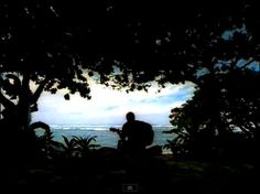 the silhouette of a man sitting under a tree on top of a hill next to the ocean