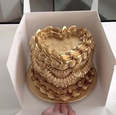 a heart shaped cake in a box with gold decorations on the top and sides, being held by a person's hand