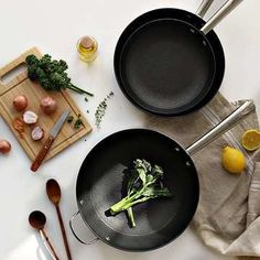 two black pans with broccoli in them next to utensils