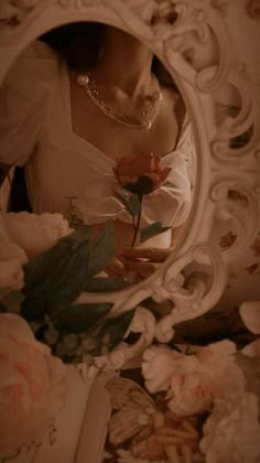 a woman in a white dress holding a flower and looking at her reflection in a mirror