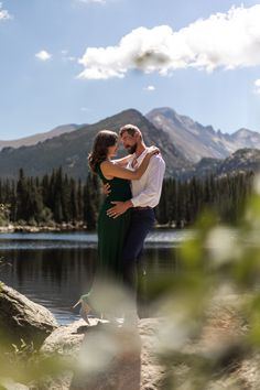 a man and woman standing next to each other near a body of water with mountains in the background