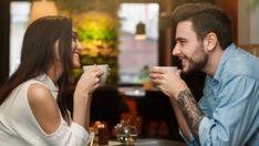 a man and woman sitting at a table with cups in their hands, talking to each other