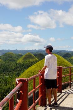 a man standing on top of a red fence next to lush green hillside covered in trees