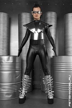 a man dressed in black and silver standing next to stacks of metal drums with his hands on his hips