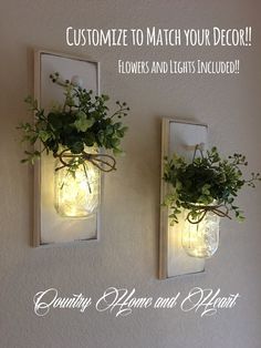 two mason jars with flowers and lights are hanging on the wall, one is filled with greenery