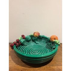 a green bowl with fruit in it sitting on a wooden table next to a white wall