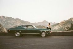 a man sitting on the hood of a car in front of mountains and hills with a woman standing next to it