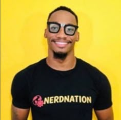 a man with glasses and a nerdnation t - shirt is standing in front of a yellow wall