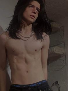 a man with long hair and no shirt standing in front of a wall wearing jeans