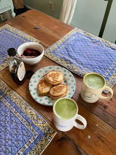 two cups of green tea sit on a table next to some pancakes and jams