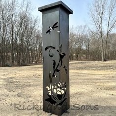 a tall metal sculpture sitting in the middle of a dirt field with trees behind it