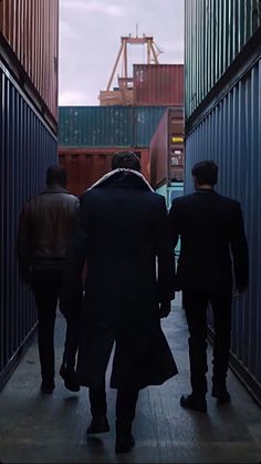 three men are walking down an alley way in front of shipping containers and one man is wearing a trench coat