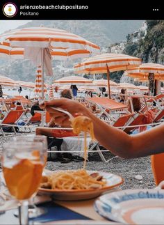 an outdoor dining area with orange umbrellas and people eating spaghetti on the beach in front of them