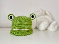 a green crocheted hat with eyes on it next to a white stuffed animal