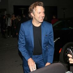 a man in a blue suit and black t - shirt smiles as he stands next to a car