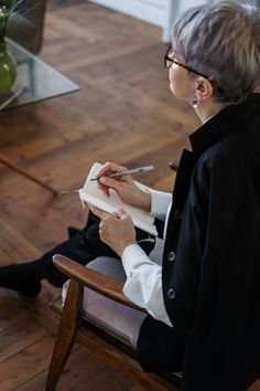 an older woman sitting in a chair writing on a piece of paper while holding a pair of scissors