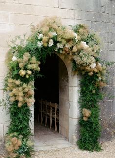 an arch covered in flowers and greenery next to a brick wall with chairs under it