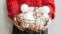 a woman holding a basket full of white and gold ornaments in her hands with a red sweater on