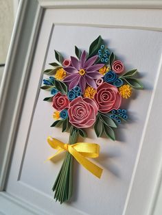 a bouquet of flowers made out of paper on a white frame with yellow ribbon tied around it