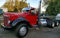 a red semi truck parked in a parking lot next to a white car and trees