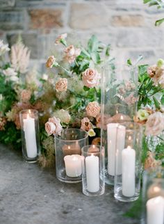 candles are lined up on the table with flowers and greenery in vases next to them