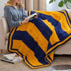 a woman sitting on a couch reading a book while holding a yellow and blue afghan