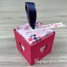 a small pink box with a blue ribbon on top and thank you sticker attached to it