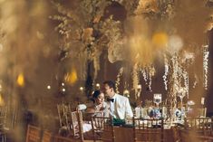 a man and woman sitting at a table with flowers hanging from the ceiling behind them