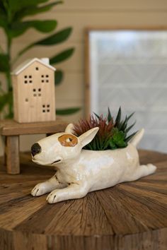 a white dog figurine sitting on top of a wooden table next to a potted plant