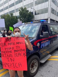 a woman holding up a sign in front of a police car that says, if you think your mask maker it hard to prevent being black in america