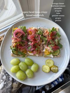 an open faced sandwich with pickles and tomatoes on a white plate next to a window