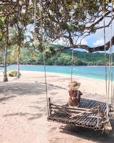 a woman sitting on a swing at the beach in front of some water and trees