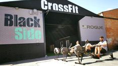 three people are riding stationary bikes in front of a black side building with the word crossfit on it