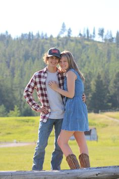 two young people standing next to each other on a wooden fence with trees in the background