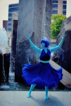 a woman with blue hair is dancing in front of a fountain