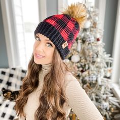 a woman wearing a red and black plaid pom - pom beanie standing in front of a christmas tree