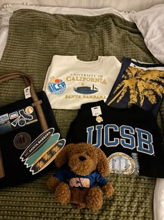 a teddy bear sitting on top of a bed next to t - shirts and a purse