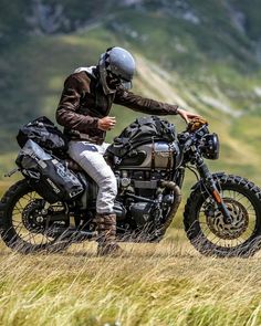 a man riding on the back of a motorcycle down a grass covered field next to a mountain