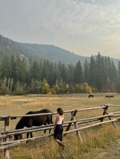 a woman standing next to a wooden fence with horses grazing in the field behind her