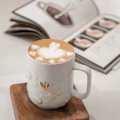 a coffee cup with a bee on it sitting on a wooden coaster next to an open book