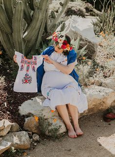 a woman sitting on the ground next to a cactus holding a white shirt with flowers in her hair