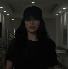 a woman with long black hair wearing a baseball cap in a dark room and looking at the camera