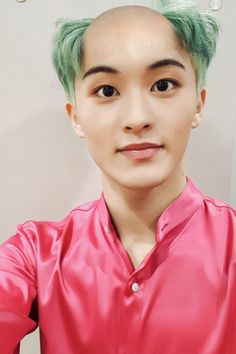 a man with green hair wearing a pink shirt and looking at the camera while standing in front of a mirror