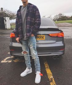 a man standing in front of a parked car wearing ripped jeans and a plaid jacket