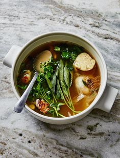 a white bowl filled with soup and vegetables on top of a stone counter next to a spoon