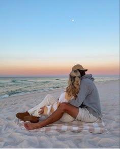 a man and woman sitting on top of a beach next to the ocean at sunset