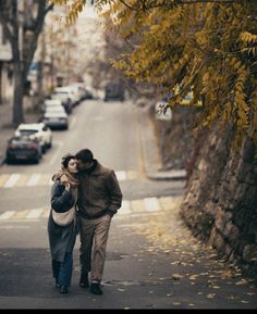 a man and woman walking down the street with cars parked on either side of them