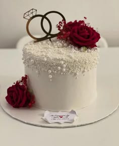 a wedding cake decorated with white frosting and red flowers on a plate next to a couple's rings