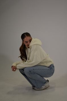 a woman squatting down in front of a white background wearing a hoodie and jeans